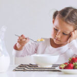 Girl looking sadly at bowl of cereal, with plate of fruit and pitcher of milk on the table.