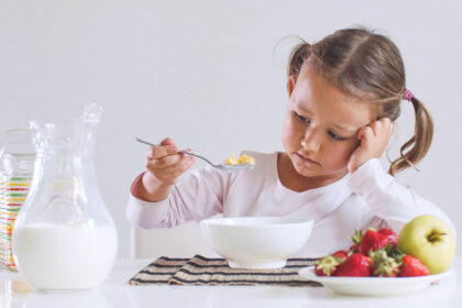 Girl looking sadly at bowl of cereal, with plate of fruit and pitcher of milk on the table.