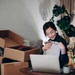 a young woman sits at her laptop with multiple boxes she received from online purchases