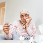 Woman sits looking dejectedly at thermometer surrounded by crumpled tissues.