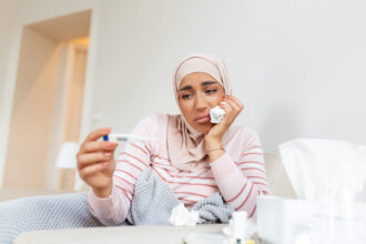 Woman sits looking dejectedly at thermometer surrounded by crumpled tissues.