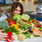 Woman embraces a big pile of assorted vegetables piled on a kitchen countertop.