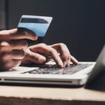 unseen person holds credit card while typing on a laptop