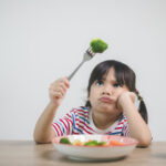 Girl sits at a table with a bowl of vegetables looking skeptically at a piece of broccoli speared on her fork.