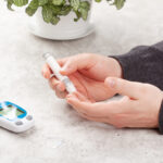 Man hands using lancet on finger to check blood ketone level. The meter is on the table nearby.