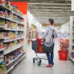 Person standing in grocery store aisle pushing a shopping cart.