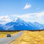 Car driving down a road with golden fields on either side, leading towards snow-capped mountains on the South Island of New Zealand