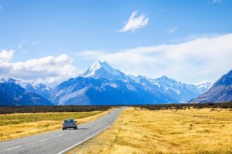 Car driving down a road with golden fields on either side, leading towards snow-capped mountains on the South Island of New Zealand