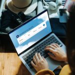 Young Asian woman shopping online for flight ticket on airline website with laptop at home, sitting next to suitcase with sun hat, camera, clothings, headphones, smartphone and passport. Getting ready for vacation. Travel and vacation concept