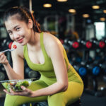 Woman in sportswear sitting on a bench in a gym eating salad and smiling into the camera.