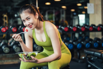 Woman in sportswear sitting on a bench in a gym eating salad and smiling into the camera.