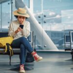 Airport Terminal: Black Woman Waits for Flight, Uses Smartphone, Receives Shockingly Bad News, Misses Flight. Upset, Sad, and Dissappointed Person Sitting in a Boarding Lounge of Airline Hub.