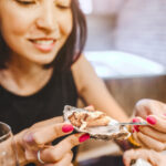 Smiling woman eating an oyster with a fork, close-up at a restaurant.