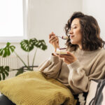 Young brunette woman sitting in a bright living savoring a bite of cheesecake.