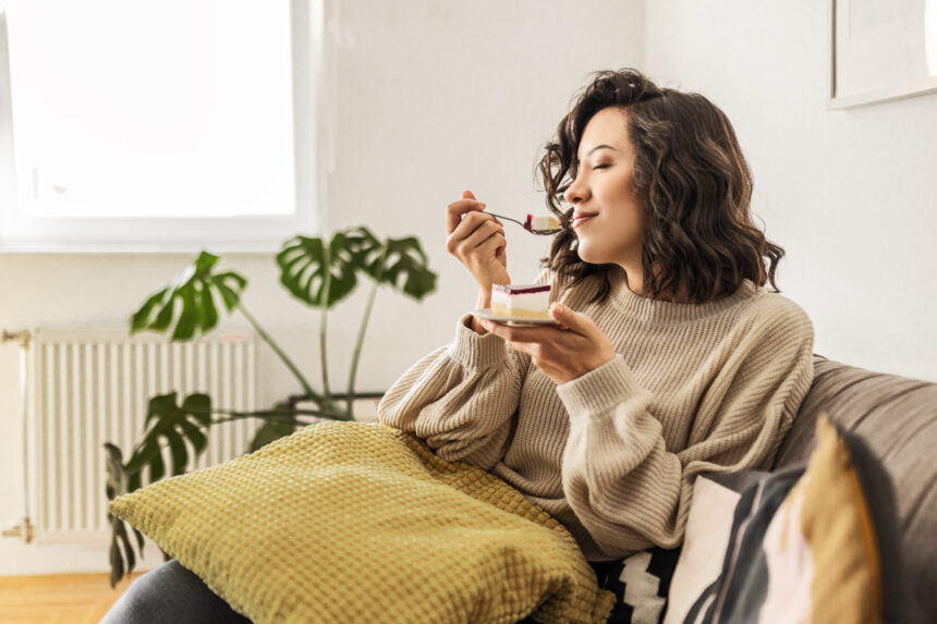 Young brunette woman sitting in a bright living savoring a bite of cheesecake.