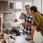 Two people in the kitchen at home. Husband is cooking for his wife, wife is tasting it.