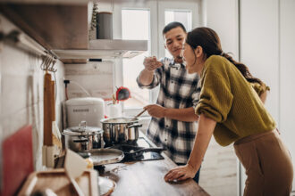 Two people in the kitchen at home. Husband is cooking for his wife, wife is tasting it.