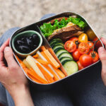 Over the shoulder view of a woman sitting on a bench holding a lunch box filled with assorted vegetables, olives, and half a sandwich.