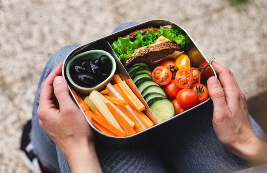 Over the shoulder view of a woman sitting on a bench holding a lunch box filled with assorted vegetables, olives, and half a sandwich.