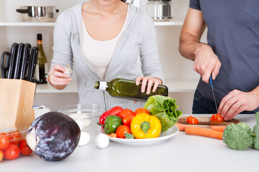 Couple cooking together with a variety of fresh bell peppers, cabbage, and other vegetables, drizzling olive oil.