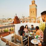 Wide shot of waiter bringing dishes to friends having rooftop dinner