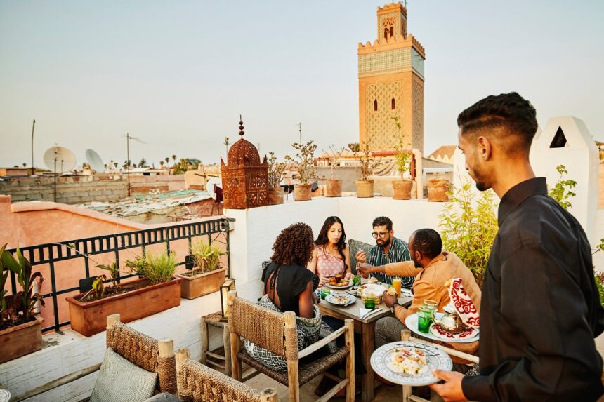 Wide shot of waiter bringing dishes to friends having rooftop dinner