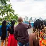 Local Tour Guide Walking and Pointing Toward the Skyline with a Group of Cheerful, Fashionable Afro-Descendant Tourists Together with a View of Panama City