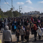 A crowd of people, some of whom carry blankets and other belongings, fills a street. Buildings are seen in the distance.