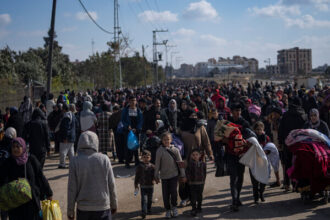 A crowd of people, some of whom carry blankets and other belongings, fills a street. Buildings are seen in the distance.