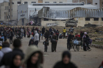 People walk on a road carrying bags, bottles of water and other items. There are tanks in the background and damaged buildings.