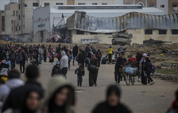 People walk on a road carrying bags, bottles of water and other items. There are tanks in the background and damaged buildings.