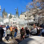 A crowd of people, some in camouflage clothing, gathers outside by a collapsed building.