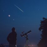 Silhouette of a man, telescope, stars, planets and shooting star under the night sky.